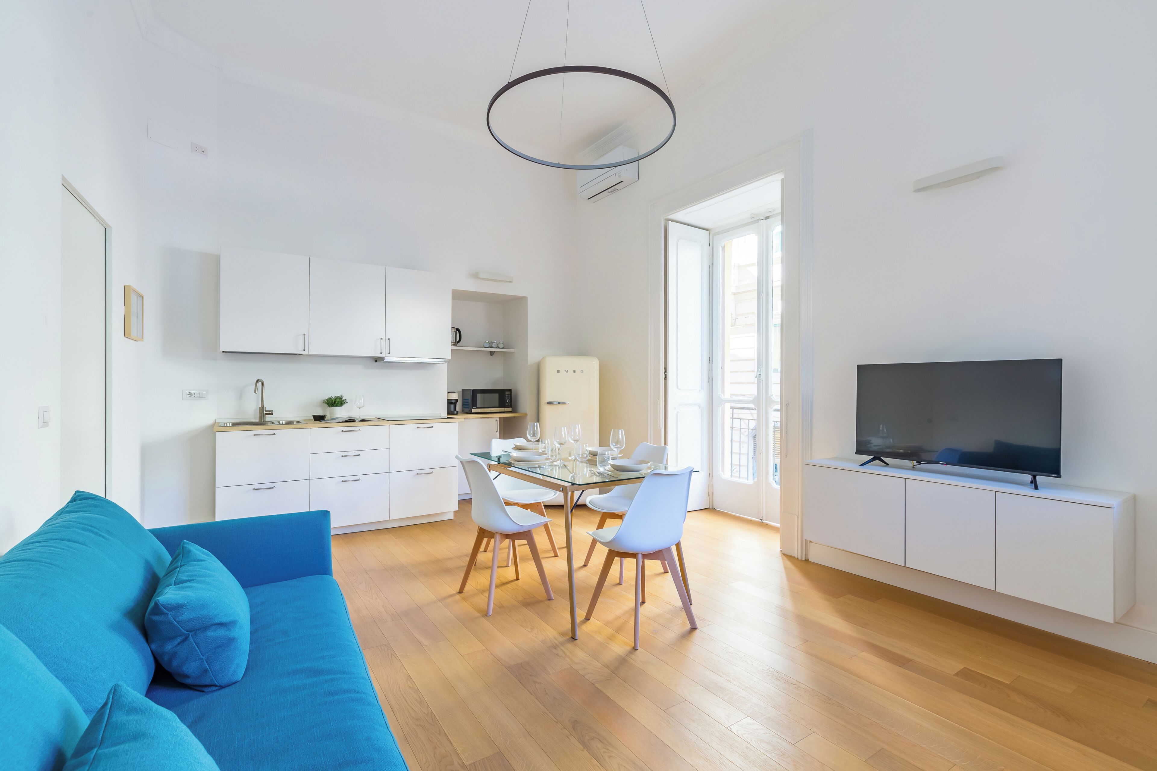 Een moderne woonkamer met een blauwe bank, een eethoek met een glazen tafel, en een keuken met witte kasten.