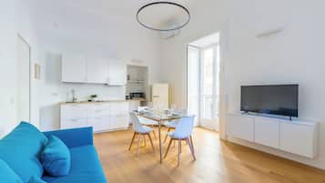 Ein modernes Wohnzimmer mit einem blauen Sofa, einem Essbereich mit einem Glastisch und einer Küche mit weißen Schränken.
