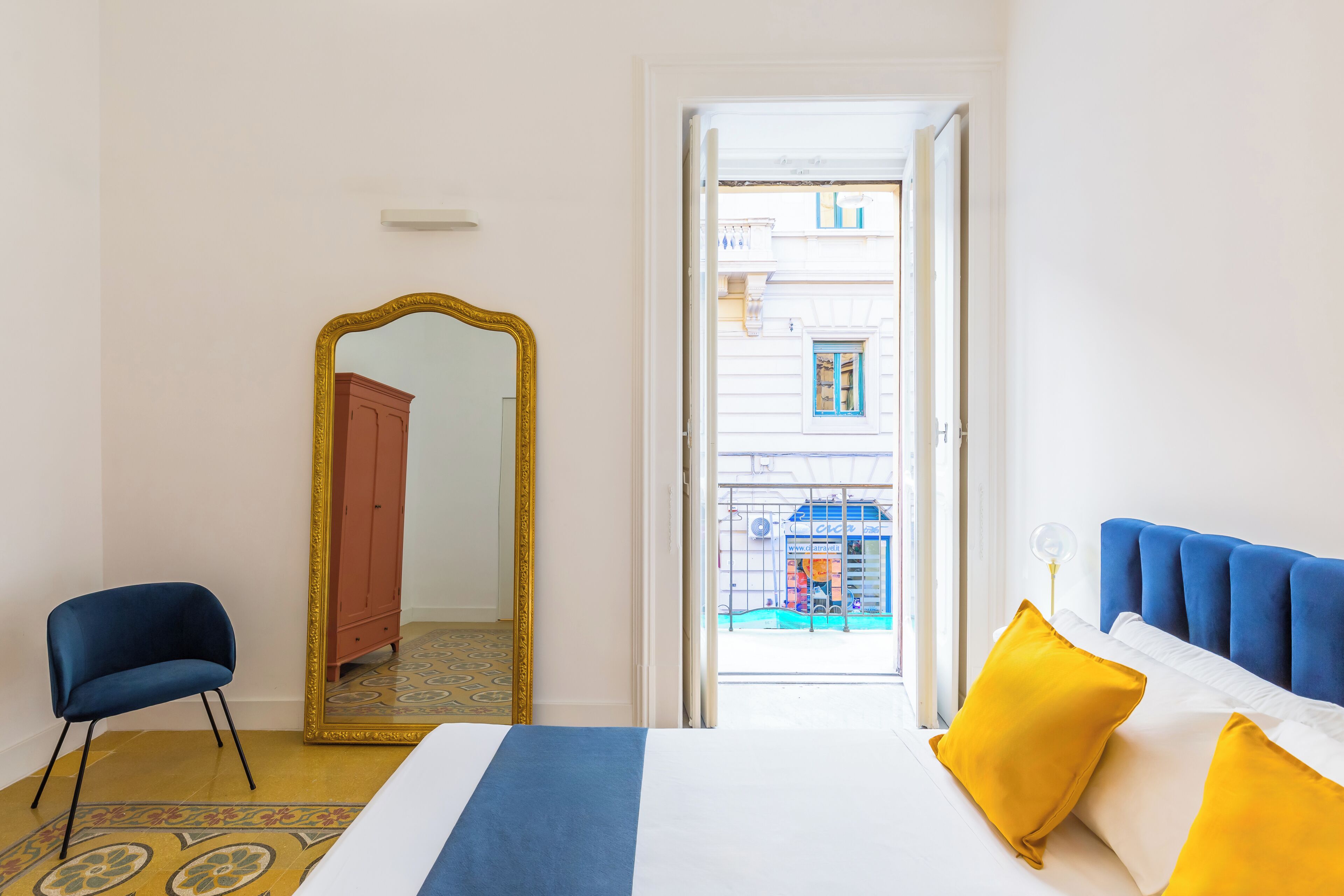 A hotel room with a bed, a chair, a large mirror, and a view of the street outside.