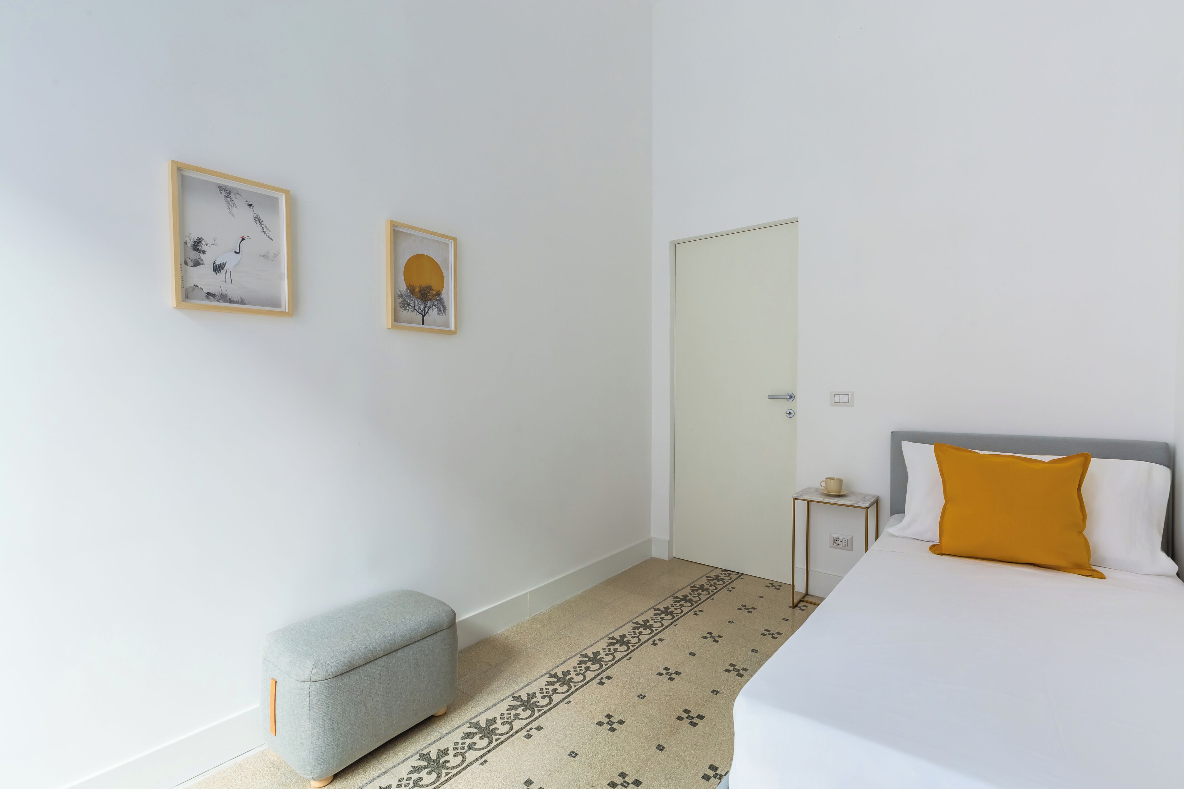 A minimalist bedroom with a bed, a small stool, a nightstand, and two framed artworks on the wall.