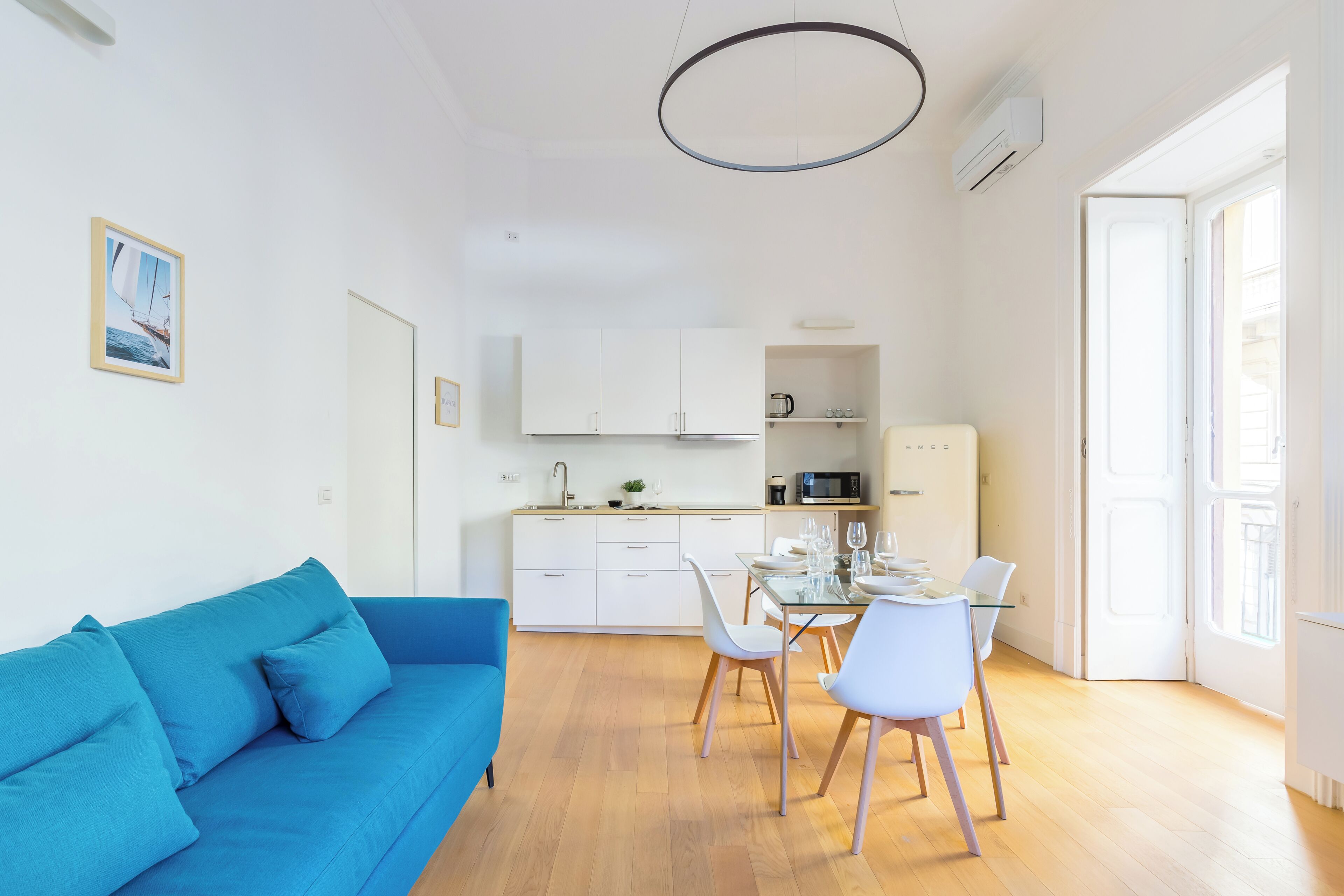 Um apartamento moderno com um sofá azul, sala de jantar com cadeiras e uma mesa posta para quatro pessoas, e cozinha com armários e eletrodomésticos brancos.