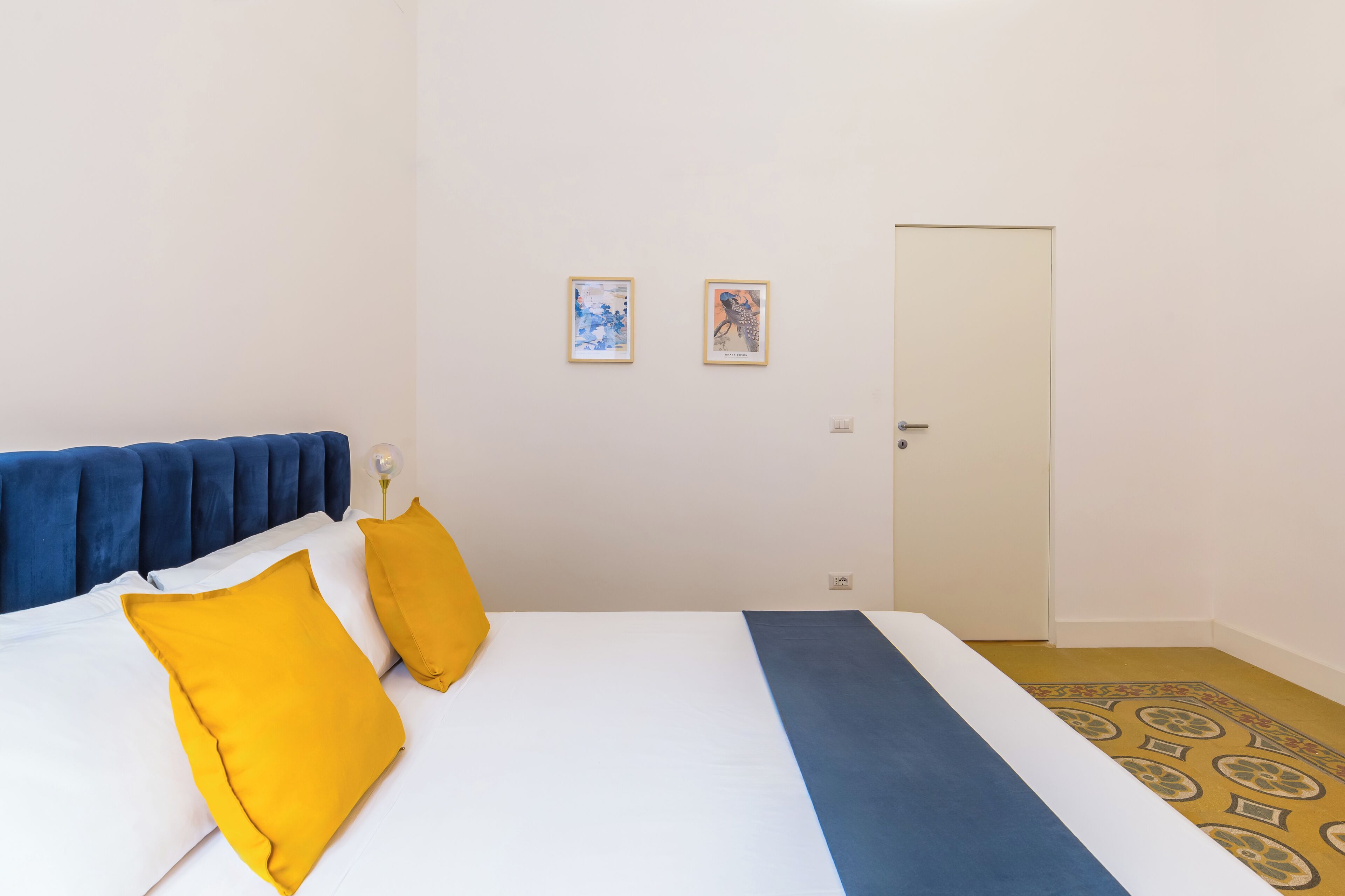A hotel room with a bed, two pillows, a bedside table, a lamp, a door, and two framed pictures on the wall.