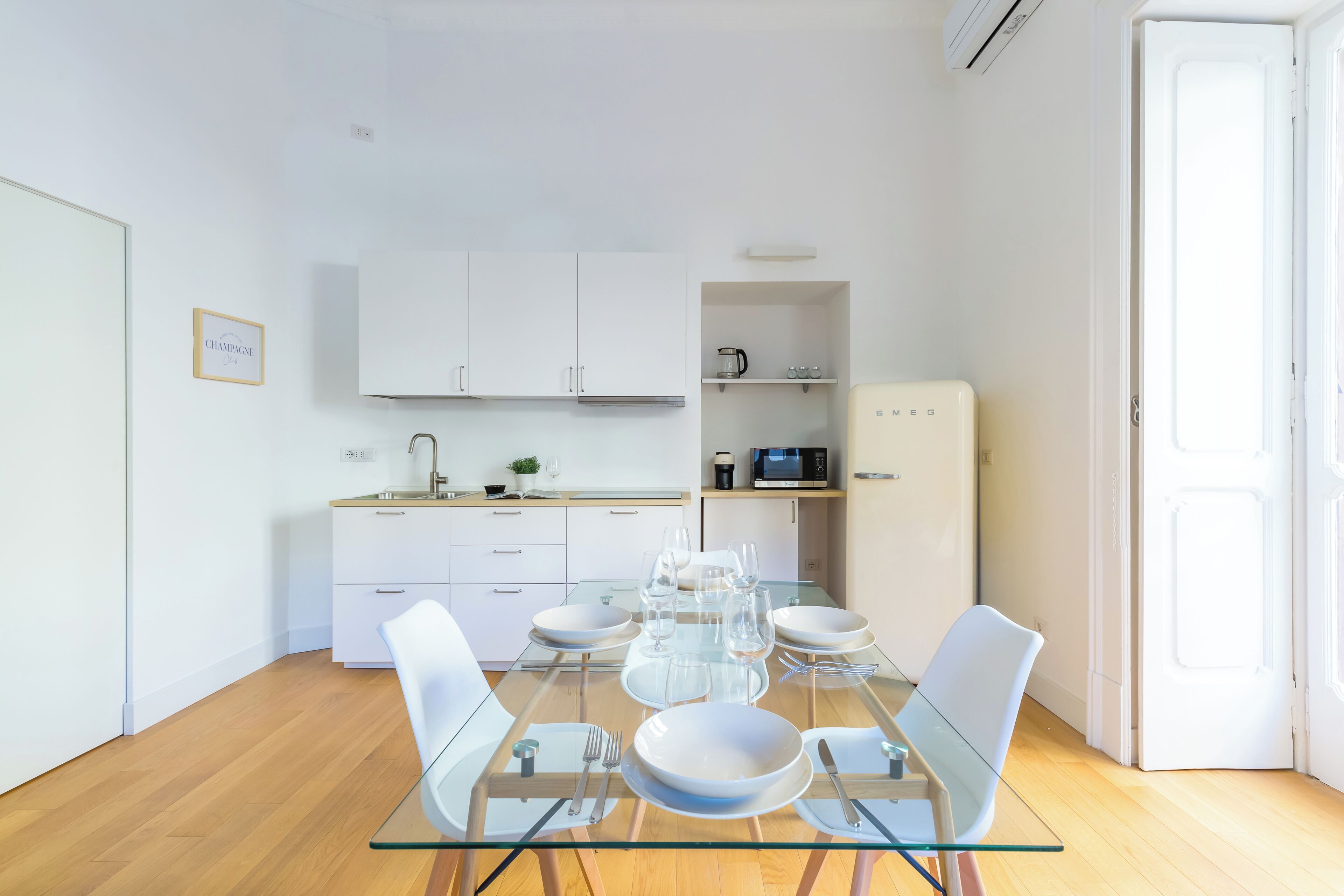 Una cocina moderna con armarios blancos, microondas y refrigerador. Hay una mesa de comedor con sillas y superficie de vidrio.