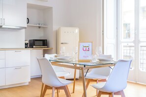 Une cuisine moderne avec des meubles blancs, un four à micro-ondes, un réfrigérateur et une table à manger en verre, assortie de chaises blanches.