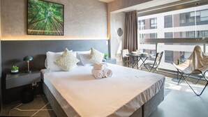 A modern hotel room with a large bed, a nightstand, a desk, and a view of the city through large windows.