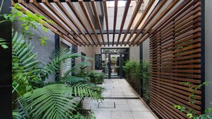 A narrow pathway flanked by lush greenery and wooden slats, leading to a modern entrance.