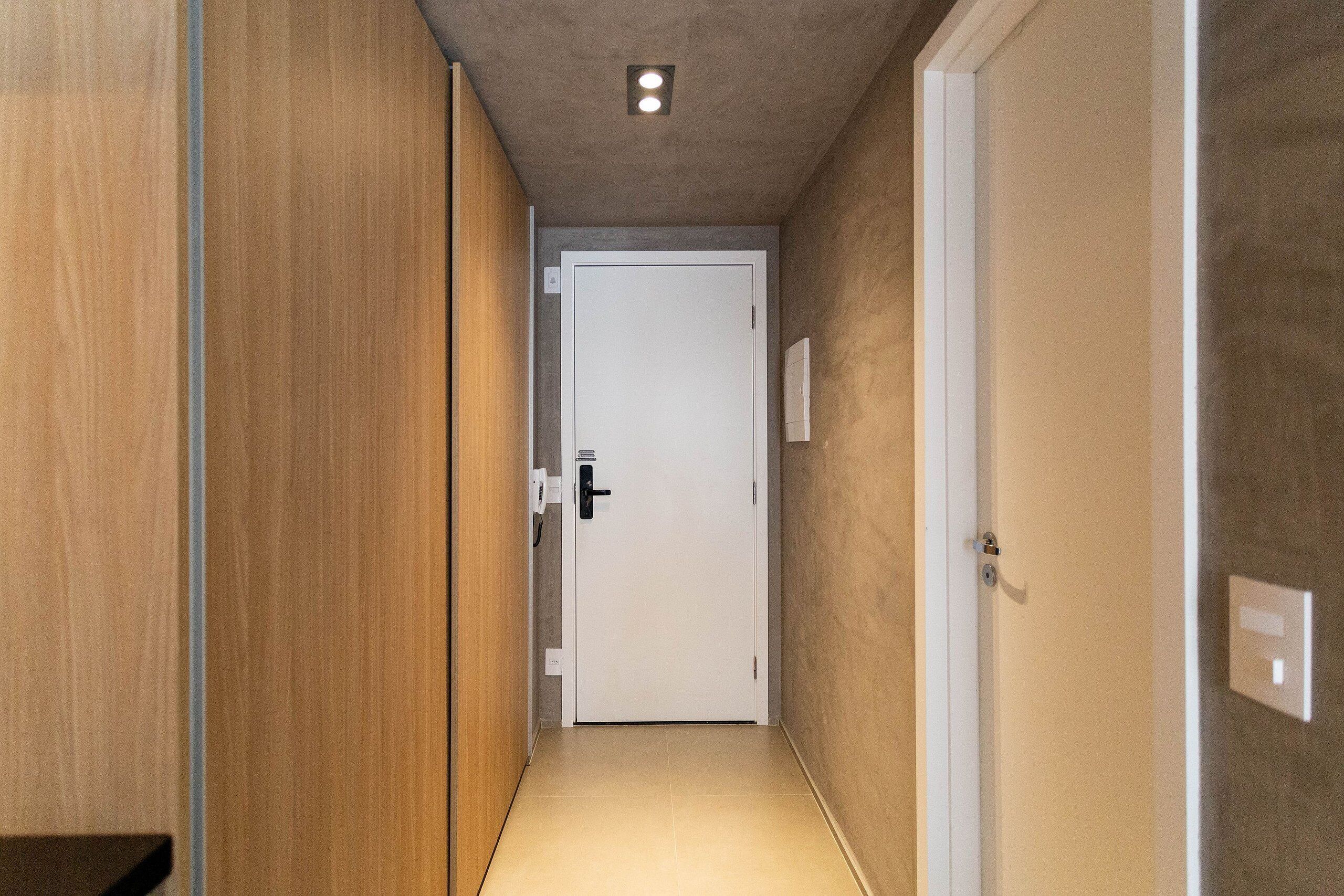 Un pasillo estrecho con una puerta de madera a la izquierda, una puerta blanca al final y una lámpara en el techo.