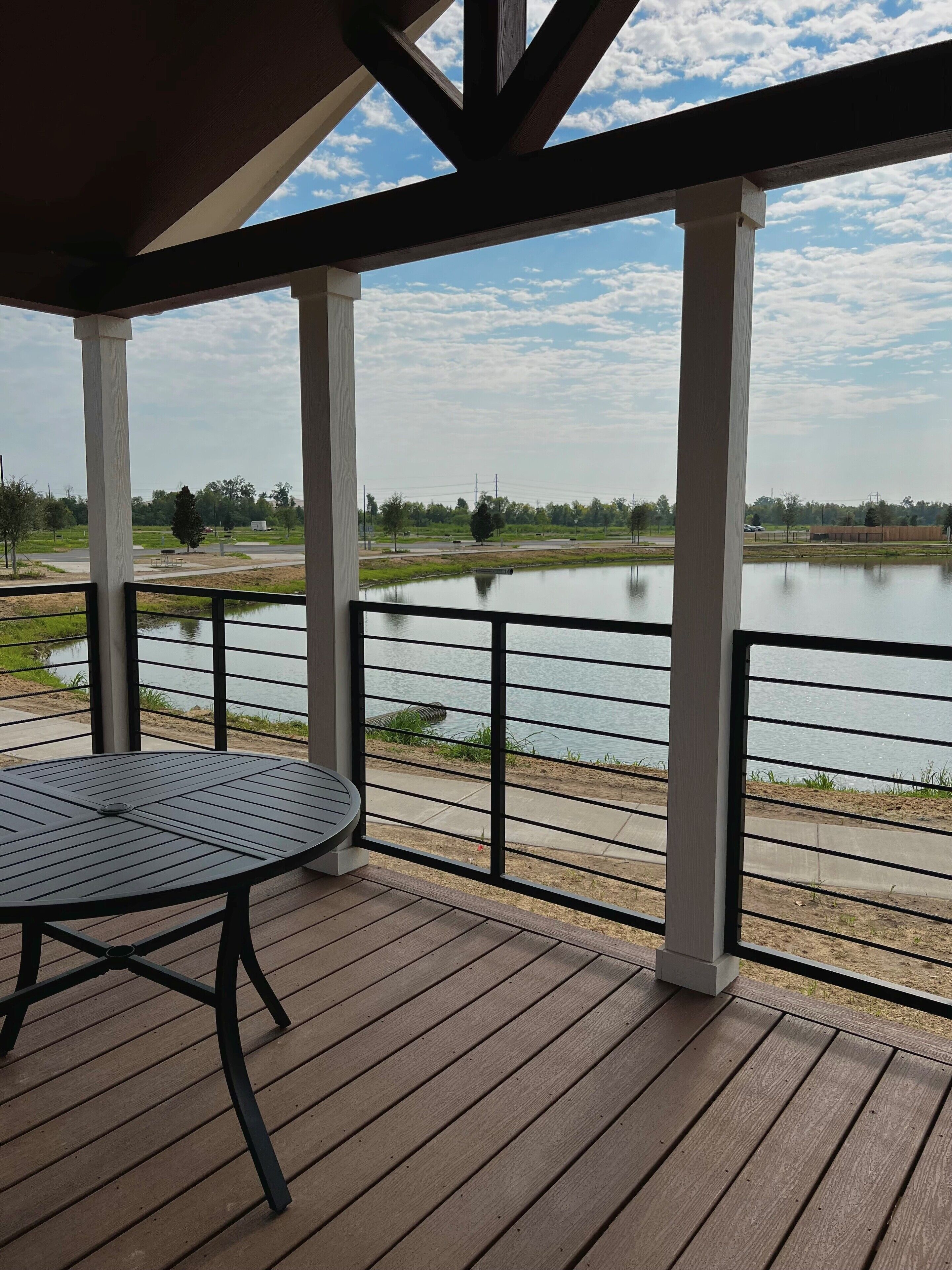 A covered outdoor area with a table and chairs, overlooking a calm body of water and a park with trees and a road.
