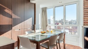 A modern dining area with a glass table, white chairs, and a city view through large windows. - Enjoybcn Fira Apartment (Barcelona)