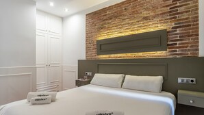 A hotel room with a large bed, a brick accent wall, white bedding, and a nightstand with a lamp. - Enjoybcn Fira Apartment (Barcelona)