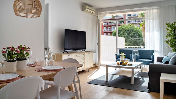 En moderne stue med spiseplads, et fladskærms-tv og en balkon med udsigt til bygninger og træer.
