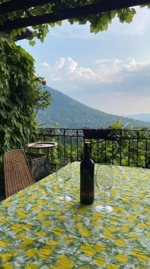 Mesa al aire libre con una botella y vasos, rodeada de follaje y un paisaje montañoso.