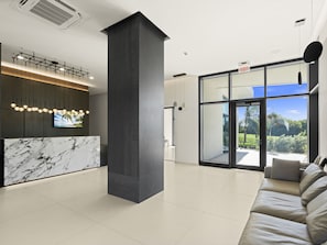 A modern lobby with a marble reception desk, a large pillar, a sofa, and a large window with a view of greenery.