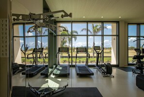 Ett gym med löpband, crosstrainers och en motionscykel, med stora fönster som ger utsikt över palmer och en golfbana.