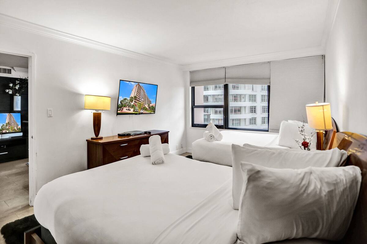 Quarto de hotel com uma cama grande, uma escrivaninha com computador, televisão e janela com vista para a cidade.