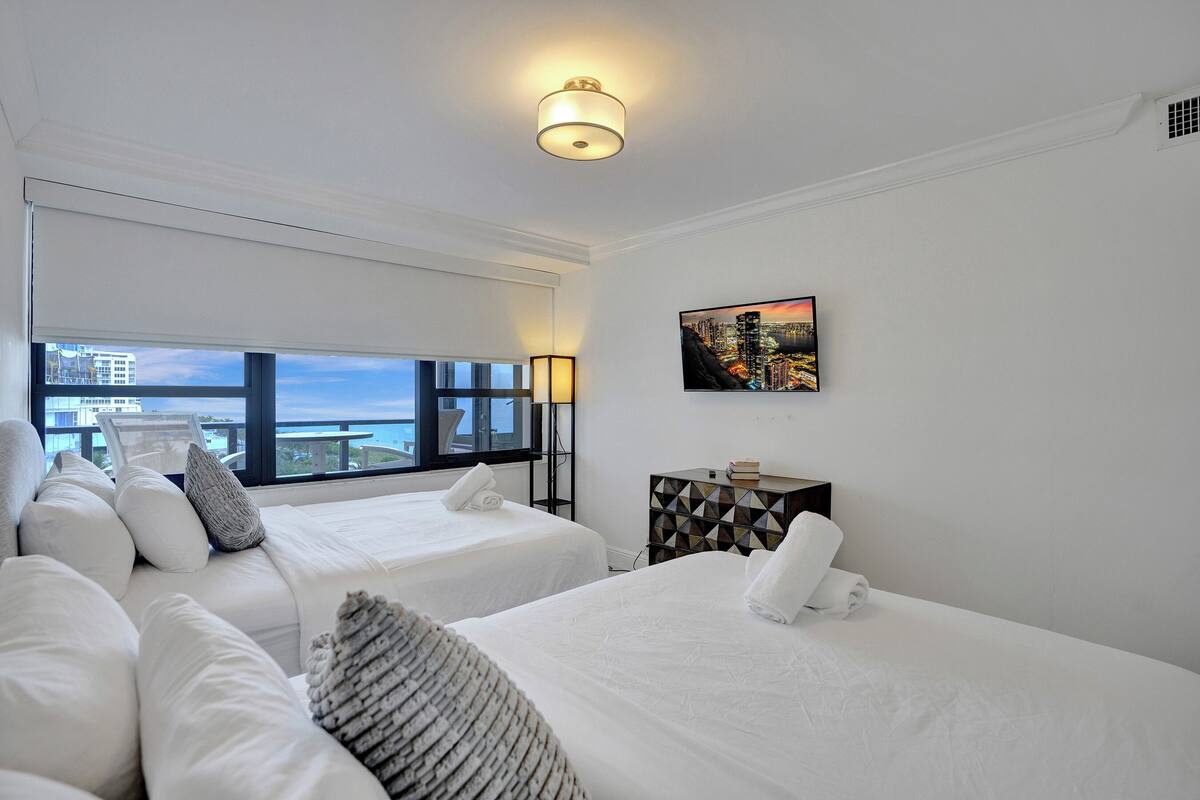 Quarto de hotel com duas camas, uma janela grande com vista para a cidade, um quadro na parede e uma mesa de cabeceira com abajur.