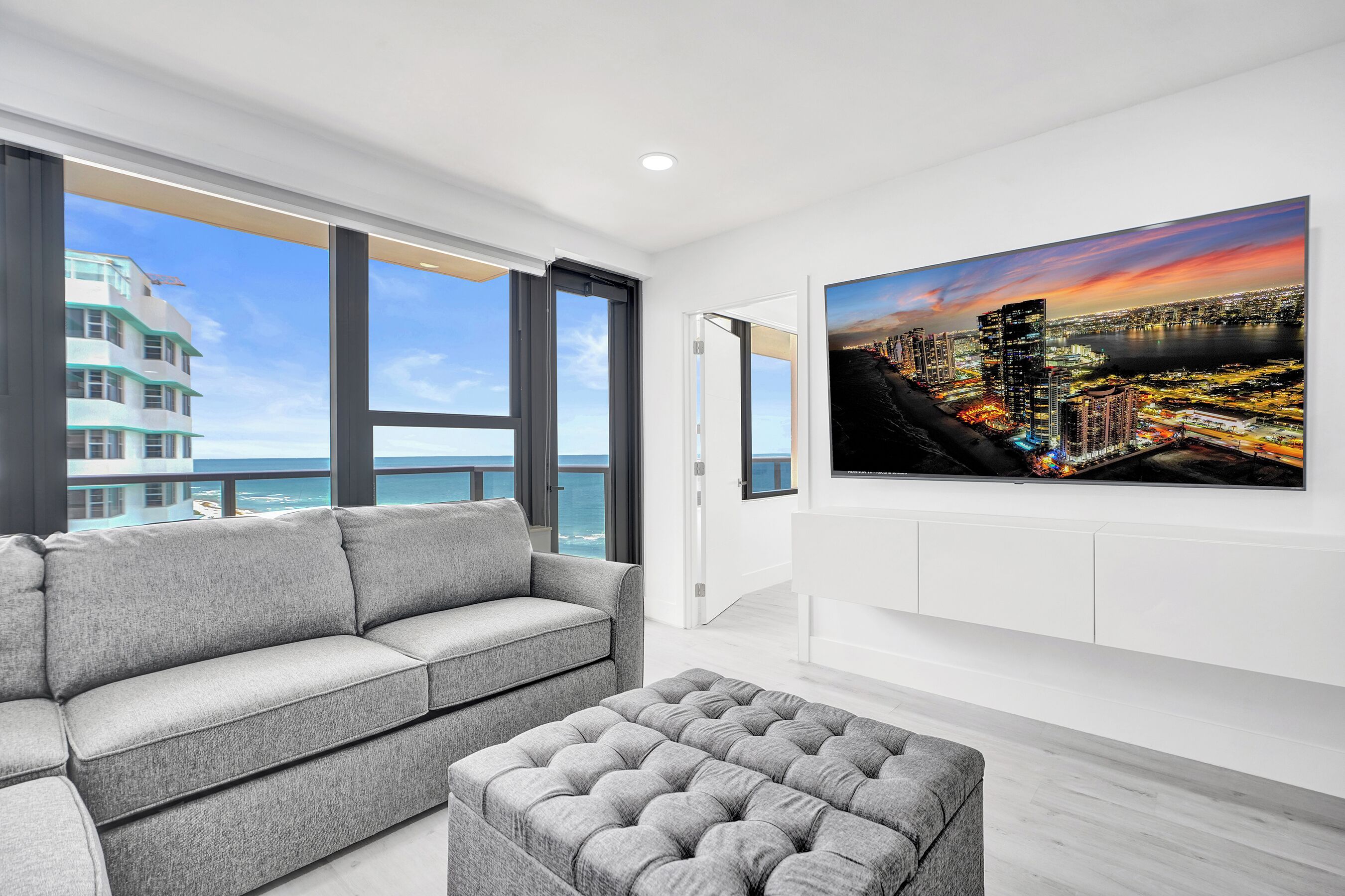 A modern living room with a grey sofa, a matching ottoman, a large cityscape painting, and a view of a high-rise building and ocean.