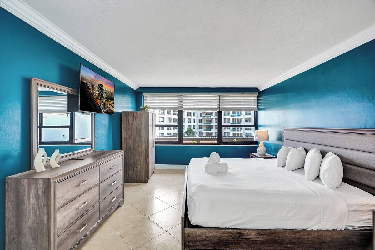 Quarto com cama, cômoda, espelho e janela com persianas.