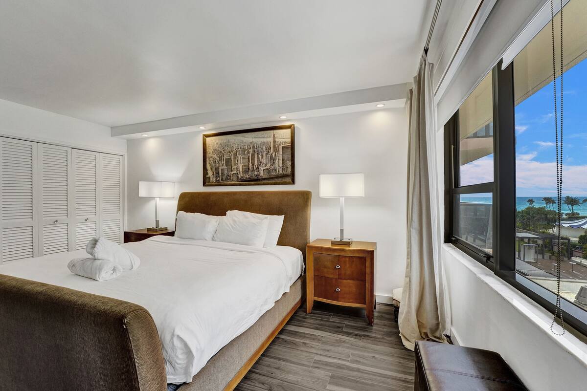 Quarto de hotel com uma cama grande, duas mesinhas de cabeceira, uma pintura de paisagem urbana e uma janela com vista.