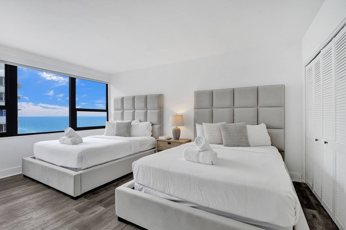 Quarto de hotel com duas camas, uma mesa de cabeceira, um abajur e vista para o oceano.