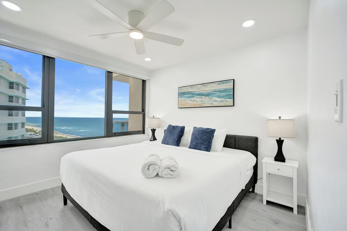 Quarto de hotel com uma cama grande, um ventilador de teto, uma mesa de cabeceira com abajur, uma pintura de uma praia e vista para o oceano.
