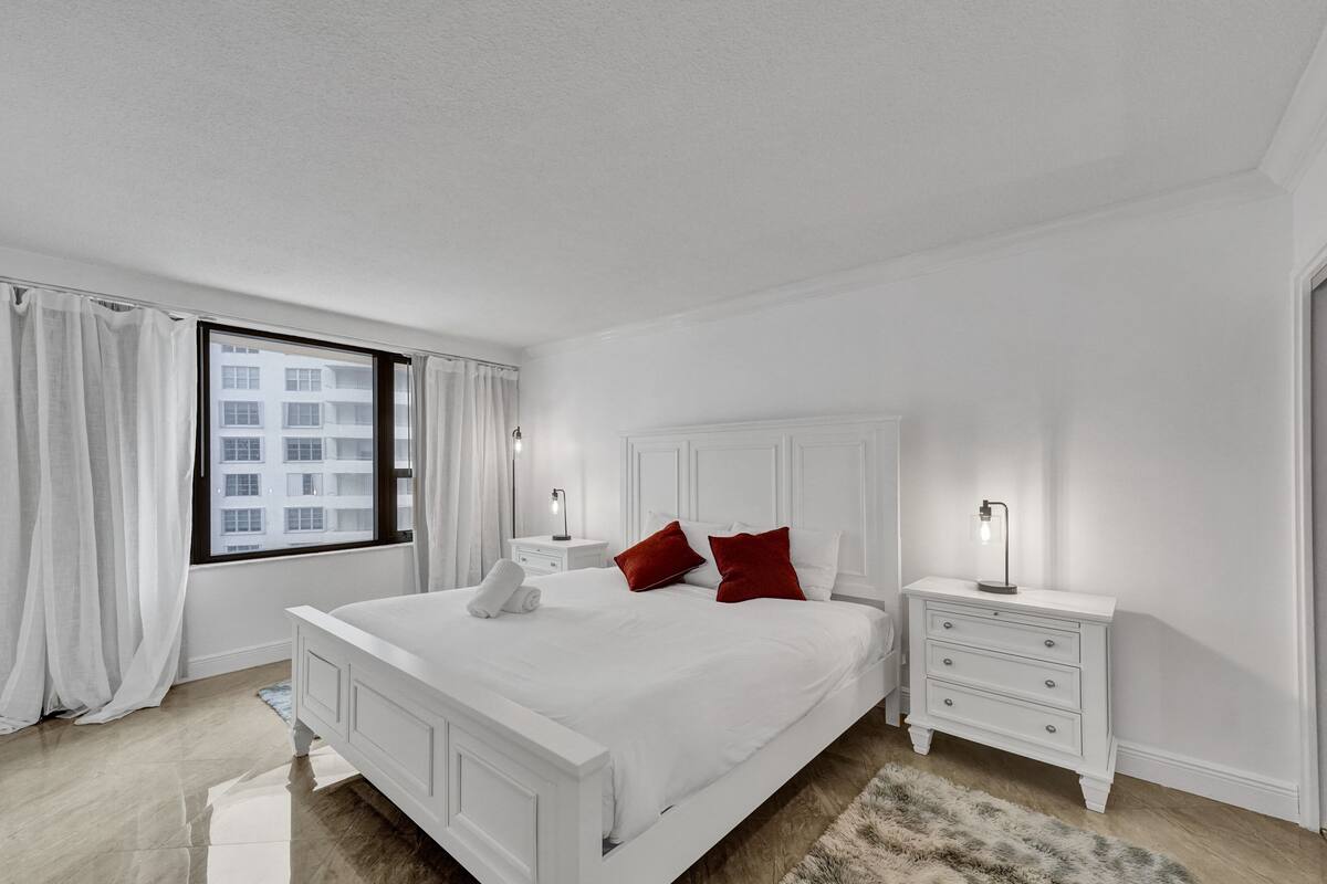 Quarto branco com uma cama grande, duas mesas de cabeceira e uma janela com cortinas.