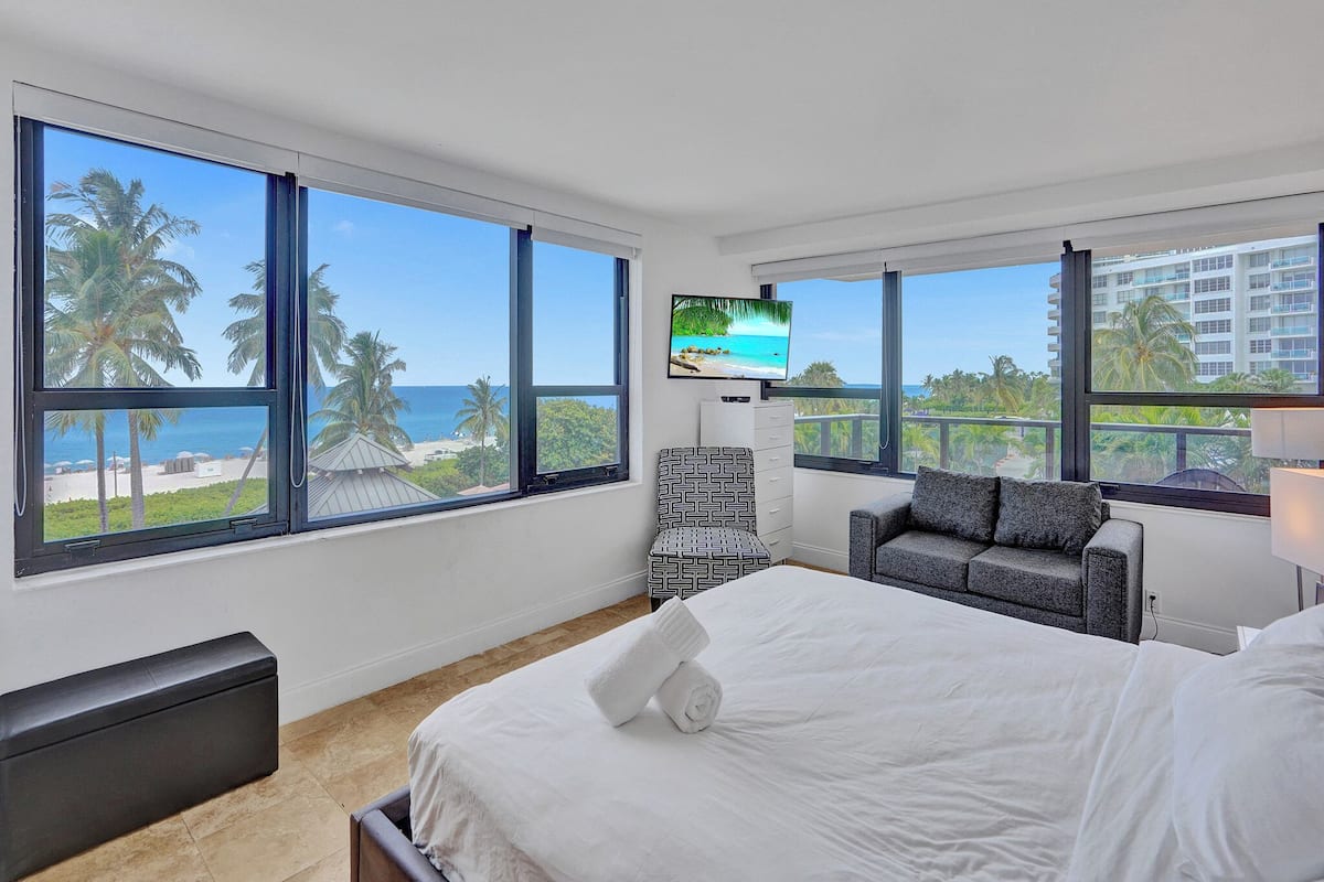 Quarto de hotel com cama, sofá, televisão e amplas janelas com vista para a praia.
