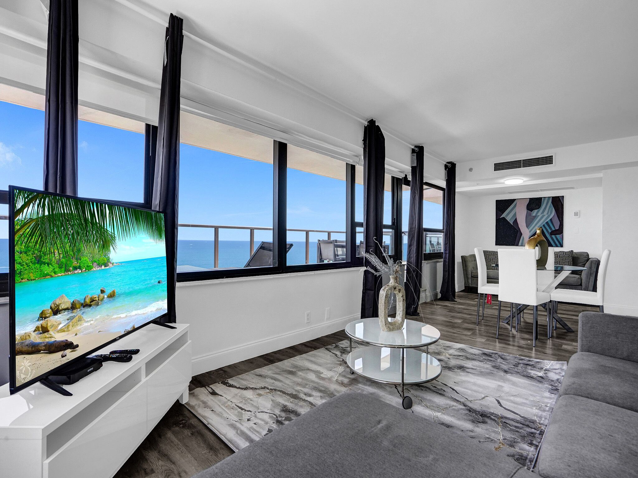Un salón moderno con un televisor grande, una mesa de centro redonda de cristal y vistas al océano a través de amplios ventanales.