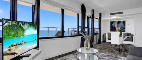 Un salón moderno con un televisor grande, una mesa de centro redonda de cristal y vistas al océano a través de amplios ventanales.