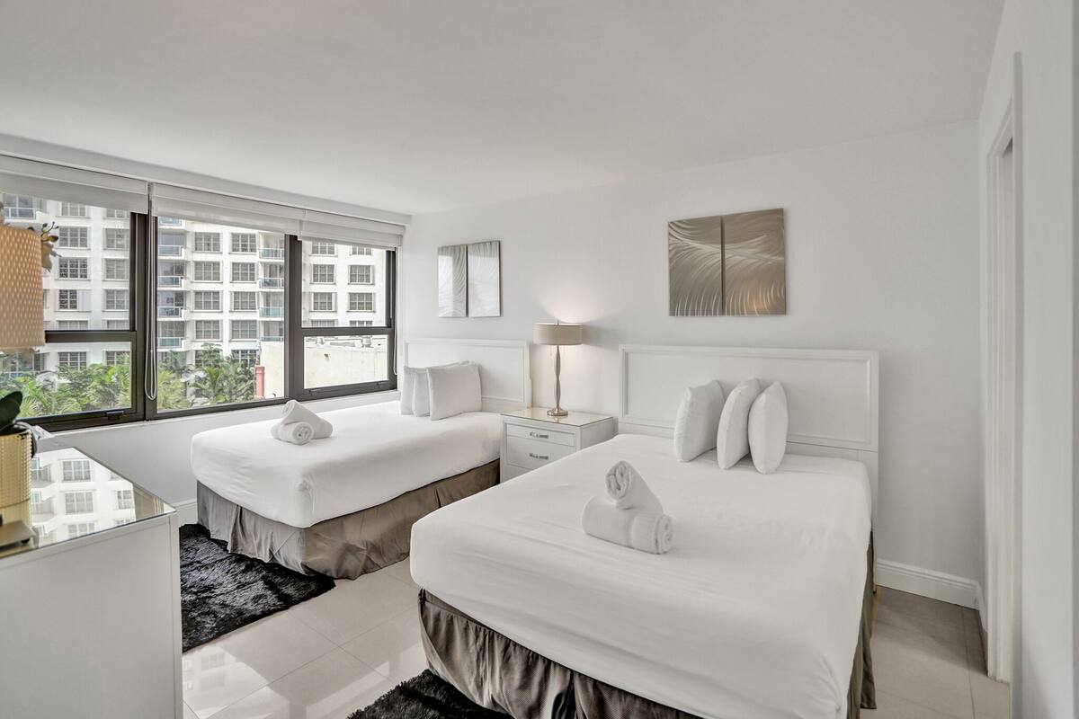 Quarto de hotel com duas camas, uma escrivaninha, uma luminária e vista para um edifício através de grandes janelas.