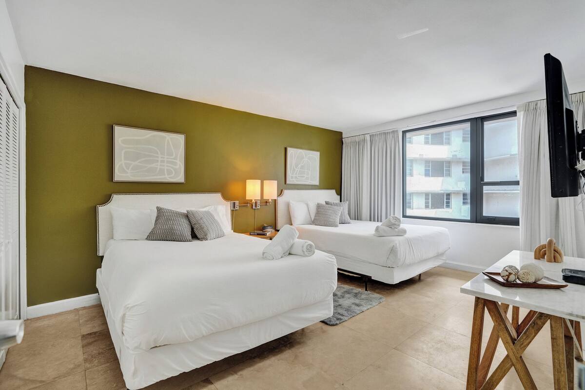 Quarto de hotel com duas camas, televisão, mesa com pão e ovos, e janela com vista para edifícios.
