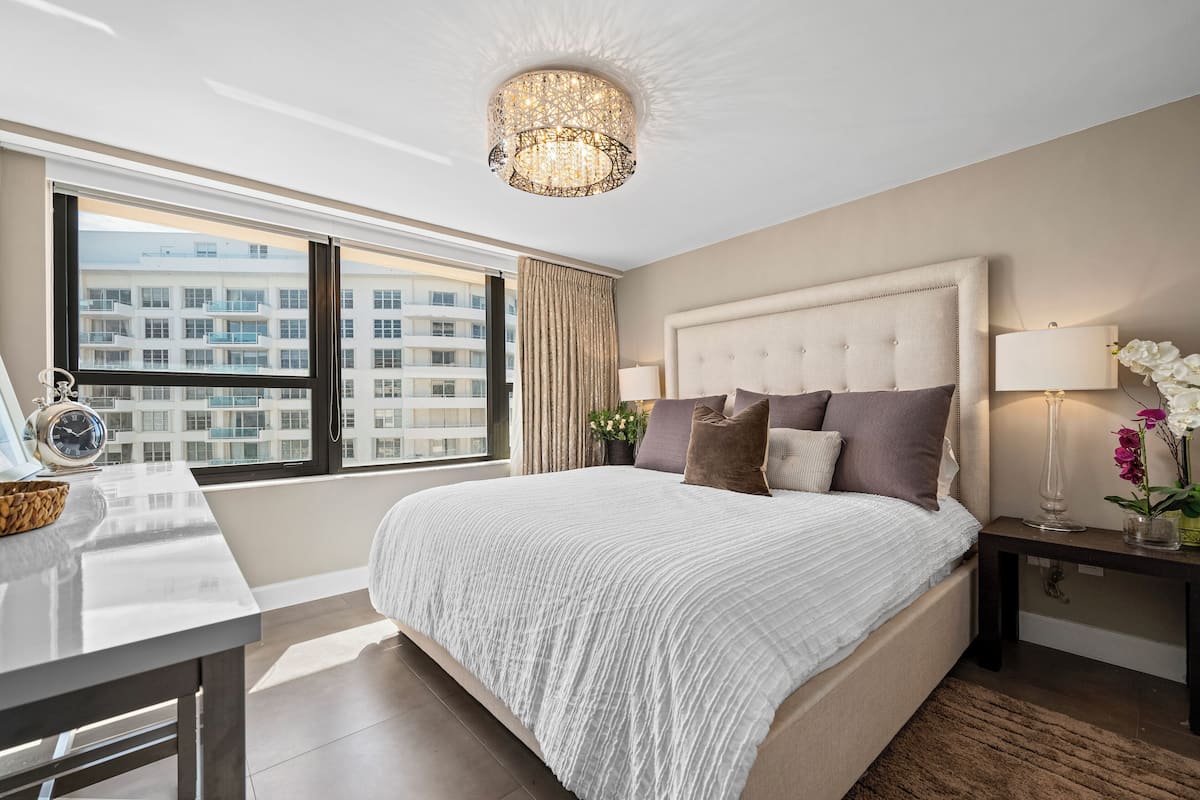 Quarto de hotel moderno com uma cama grande, uma escrivaninha, um candelabro e vista para um edifício pela janela.