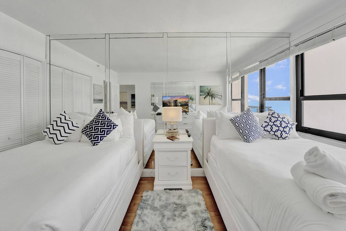 Quarto de hotel moderno com duas camas, uma mesa de cabeceira central, uma janela grande e vista para a paisagem urbana.