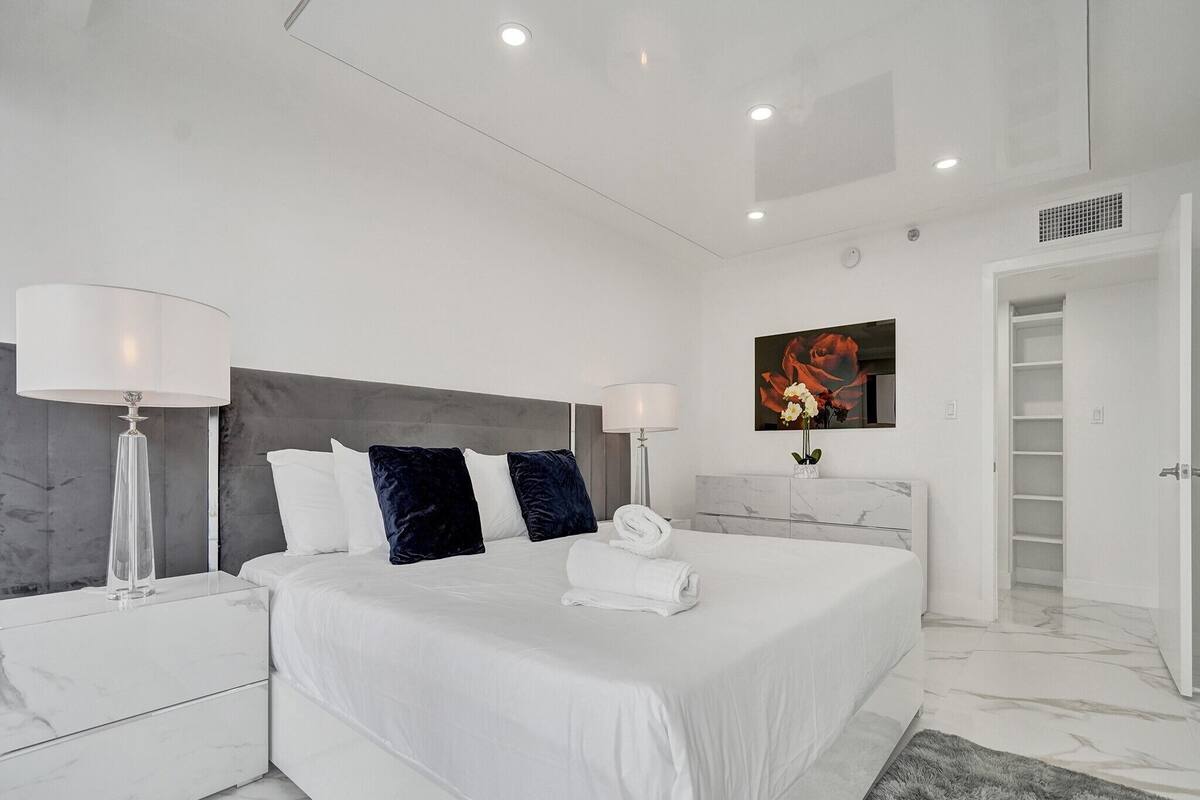 Quarto moderno com cama grande, roupa de cama branca, cabeceira de mármore, duas mesas de cabeceira com luminárias, uma mesa de apoio de mármore, um tapete cinzento e um banheiro com box de vidro.