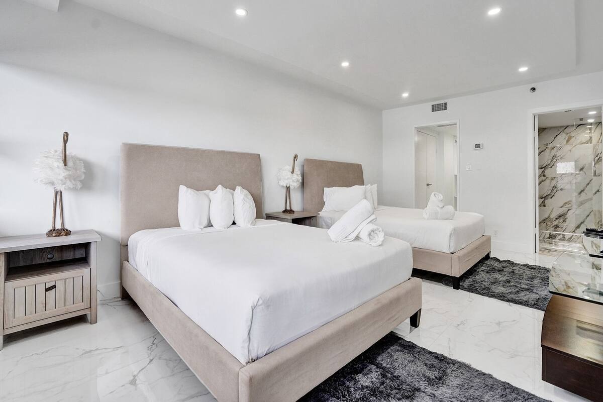 Quarto de hotel moderno com uma cama grande, uma cama menor, uma mesinha de cabeceira, um espelho e piso de mármore.
