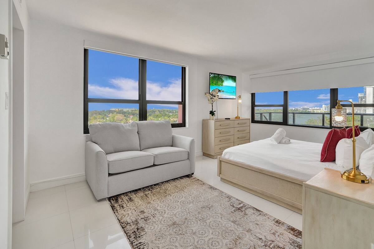 Um quarto moderno com uma cama grande, um sofá cinzento, um aparador de madeira e uma TV na parede.