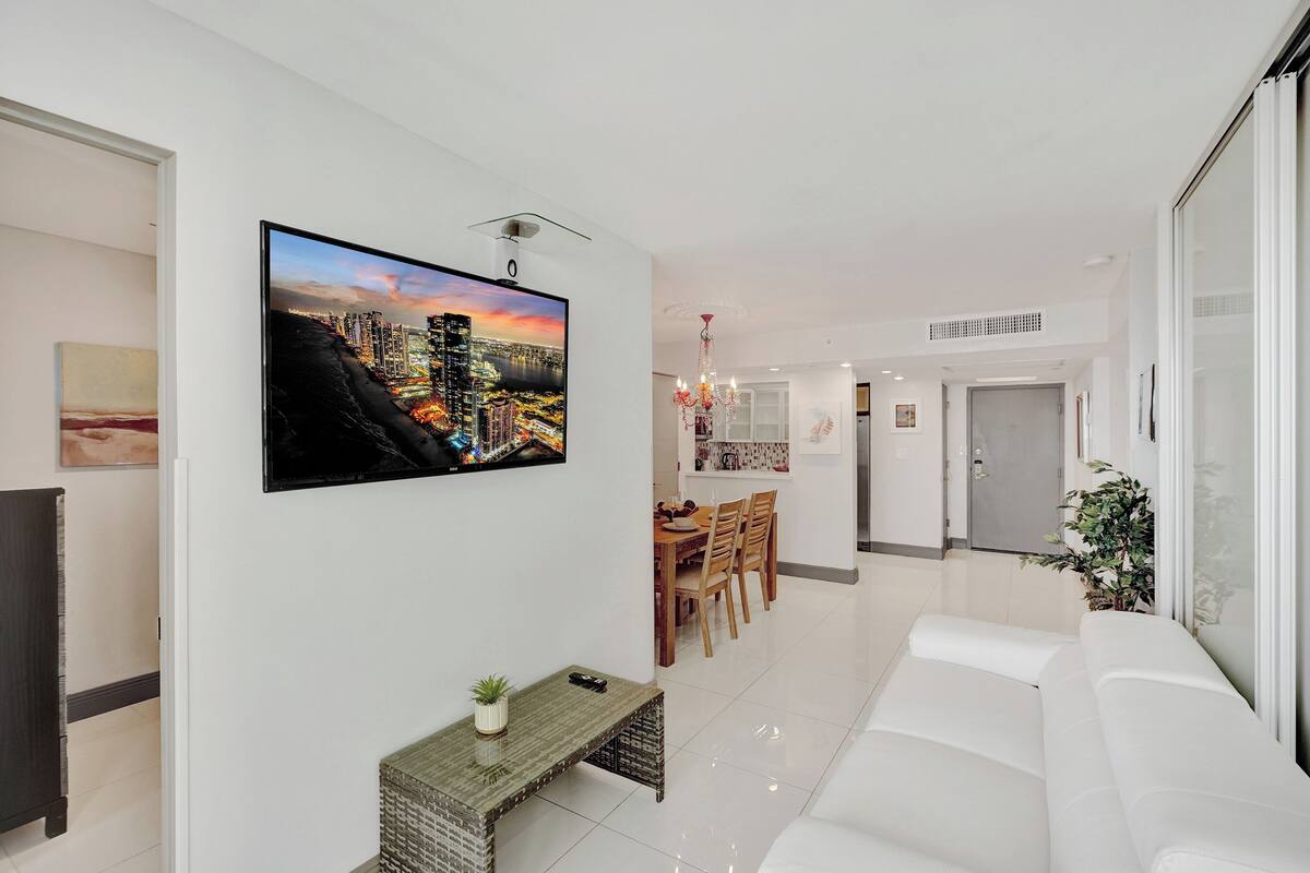 Uma sala de estar moderna com uma televisão de tela grande, um sofá branco, uma área de jantar com cadeiras de madeira e uma cozinha ao fundo.