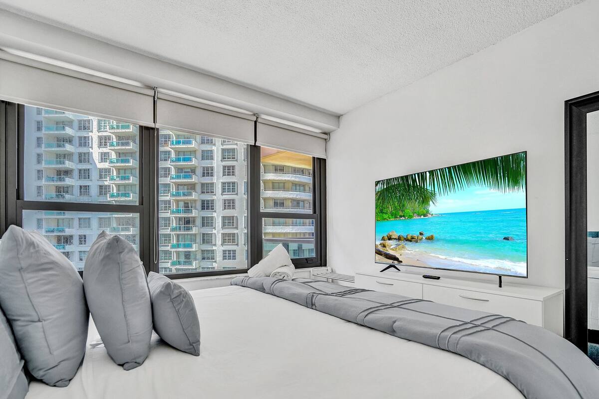 Quarto moderno com uma cama grande, televisão de tela plana e vista para um edifício alto.