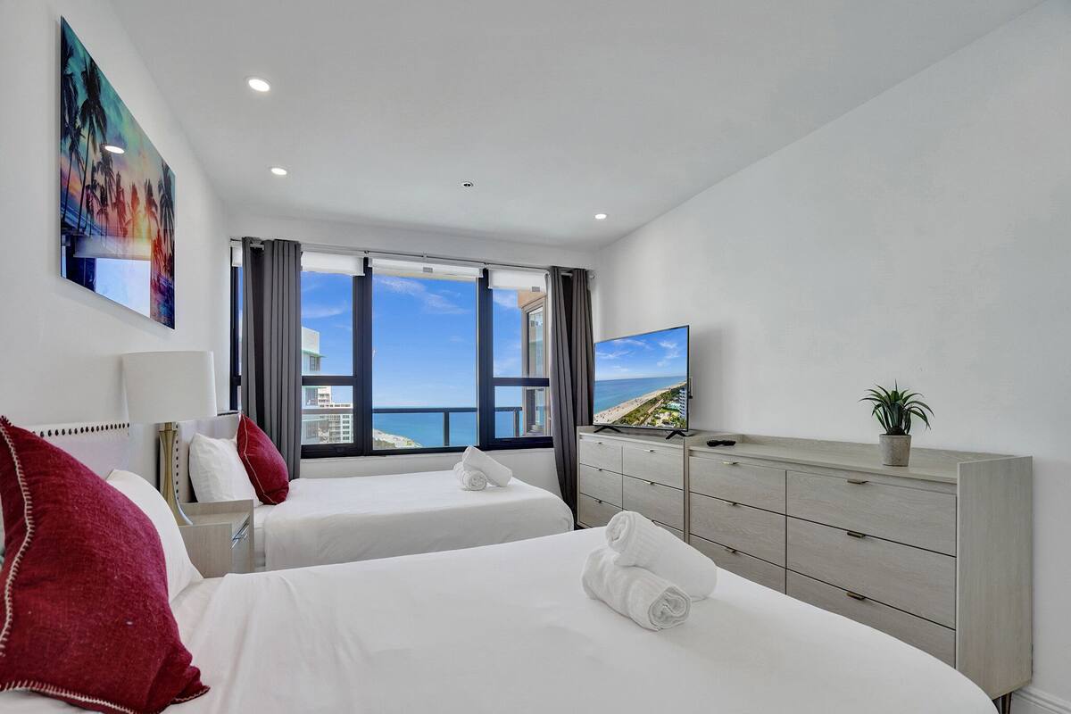 Quarto de hotel com duas camas, televisão, penteadeira e uma janela grande com vista para a praia.