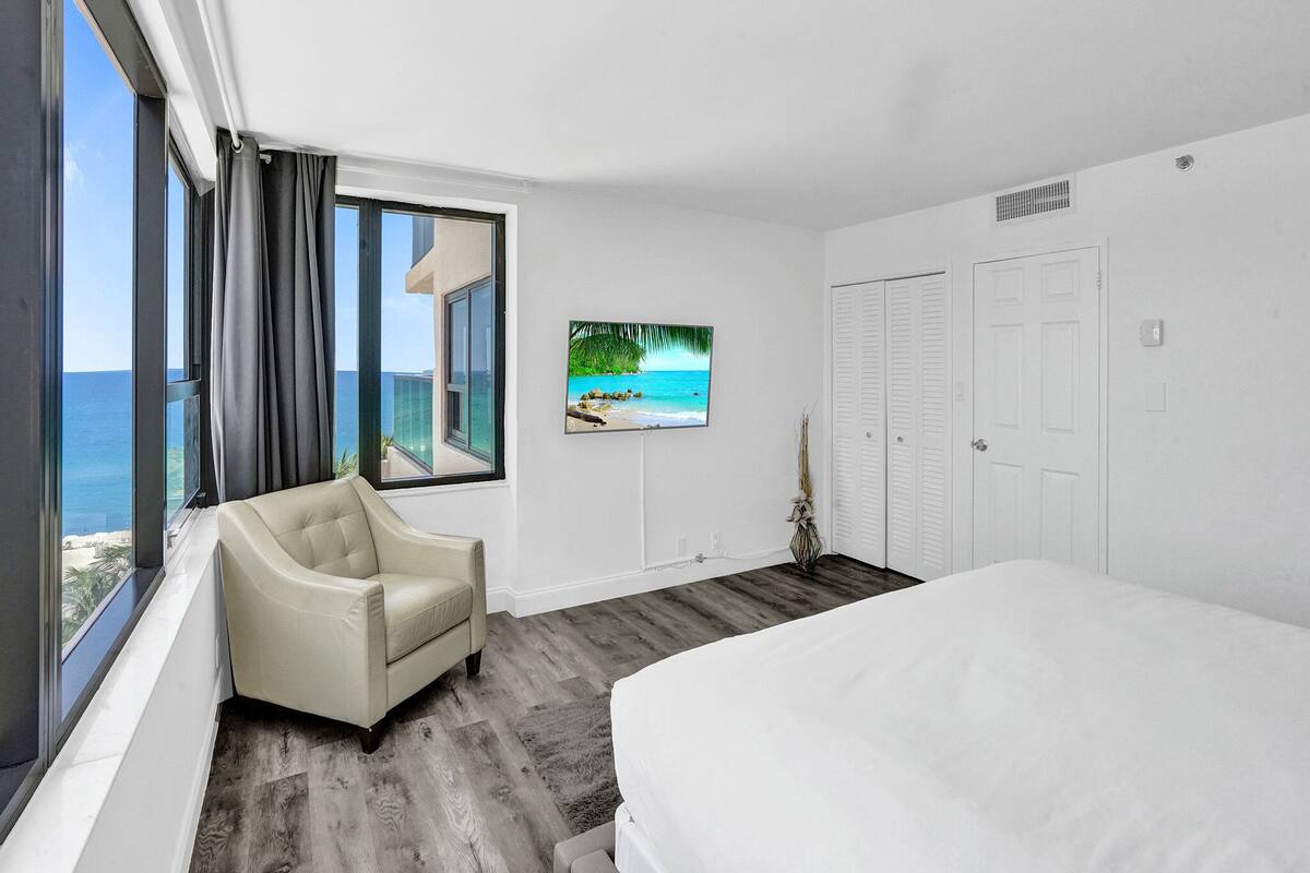 Quarto de hotel com uma cama grande, uma cadeira, uma televisão na parede e vista para o oceano.