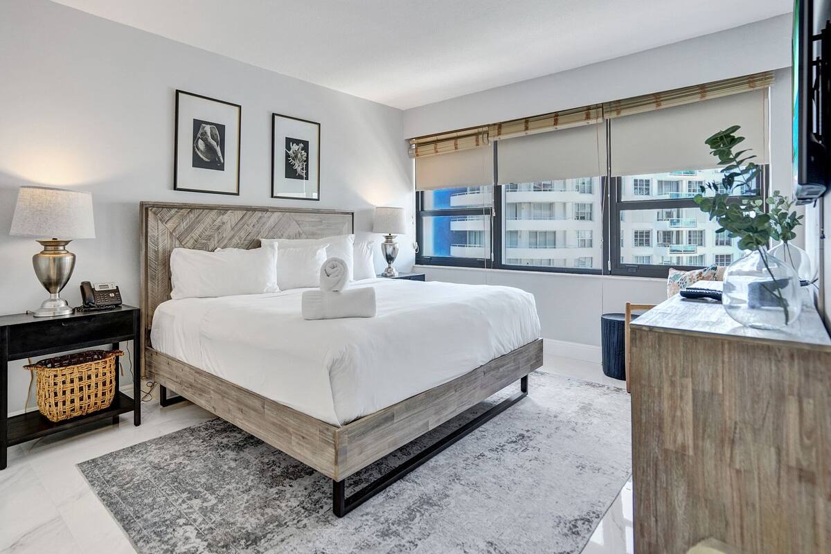 Quarto de hotel moderno com uma cama grande, uma mesa de cabeceira com luminária, uma mesinha lateral, uma escrivaninha com cadeira e uma janela com vista para edifícios.
