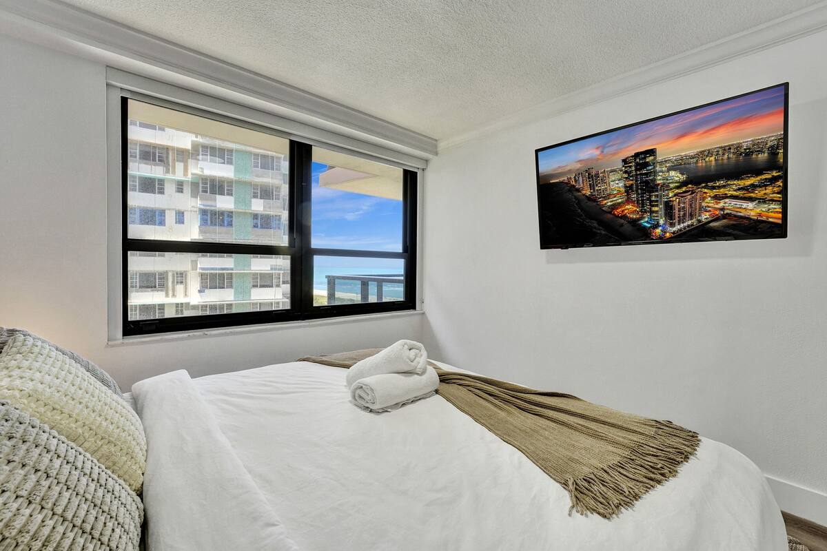 Quarto de hotel com uma grande janela, cama com roupa branca, manta bege e uma pintura da paisagem urbana na parede.