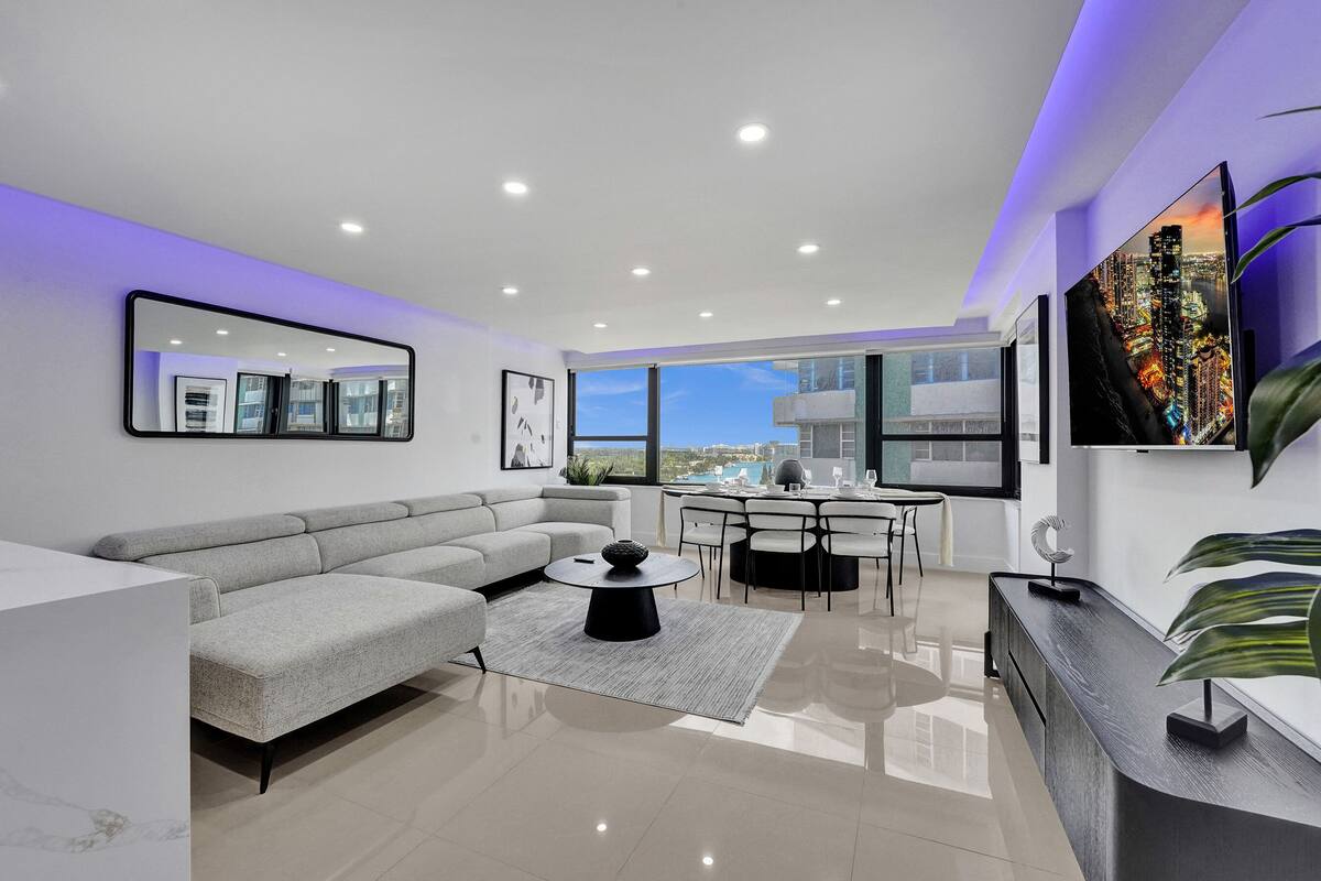 Uma sala de estar moderna com um sofá sectional cinza, uma mesa de centro redonda e uma área de jantar com uma mesa e cadeiras brancas.