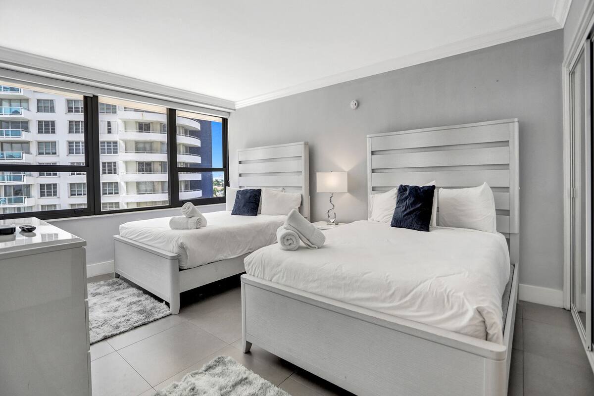 Quarto de hotel moderno com duas camas, uma escrivaninha e uma grande janela com vista para a cidade.