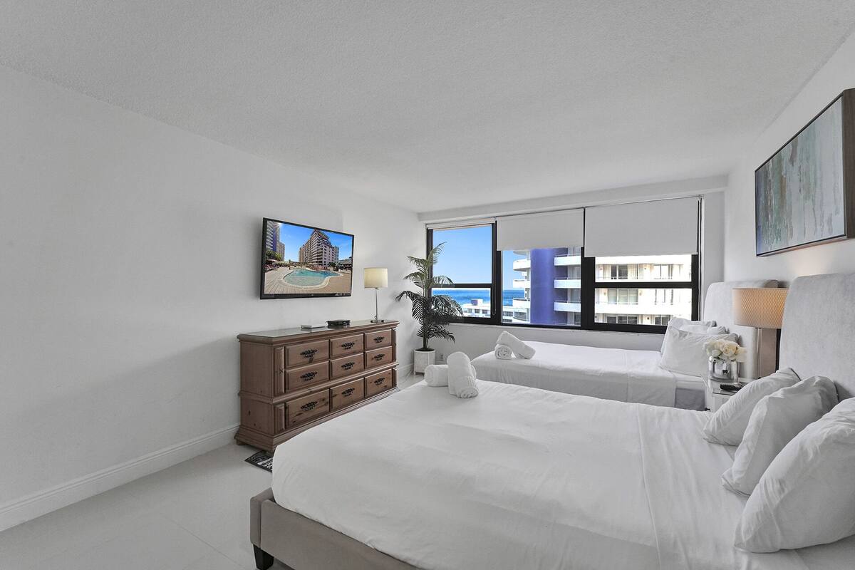 Quarto de hotel com duas camas, uma televisão na parede, penteadeira e janela com vista para edifícios e o mar.