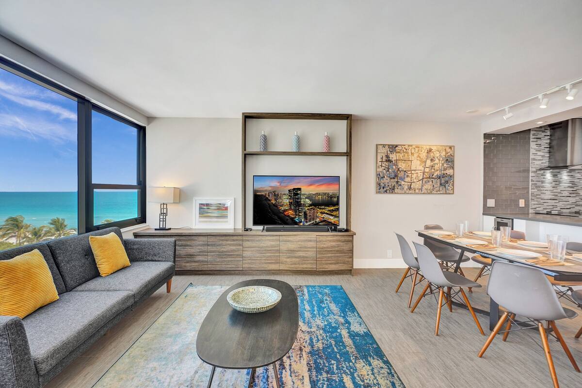 Uma sala de estar moderna com uma grande janela com vista para o oceano, um sofá cinza com almofadas amarelas, uma mesa de jantar com cadeiras e uma área de cozinha com painel de tijolos.