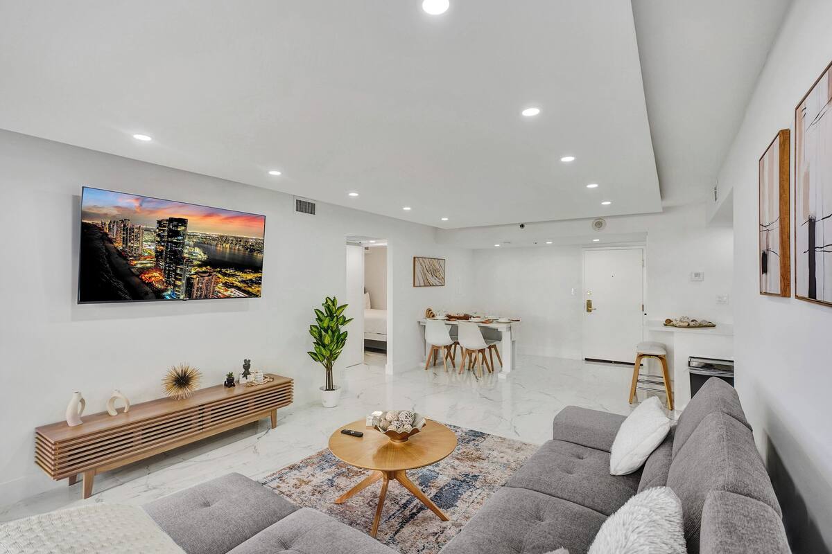 Uma sala de estar moderna com uma grande TV de tela plana, um sofá cinza, uma mesa de centro de madeira e uma área de jantar ao fundo.