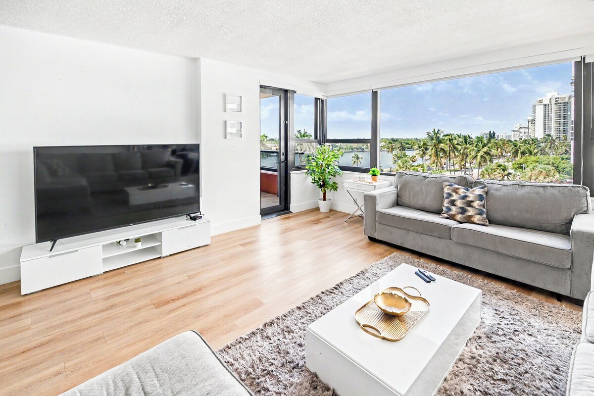 A modern living room with a flat-screen TV, grey sofa, and a view of a cityscape through large windows.