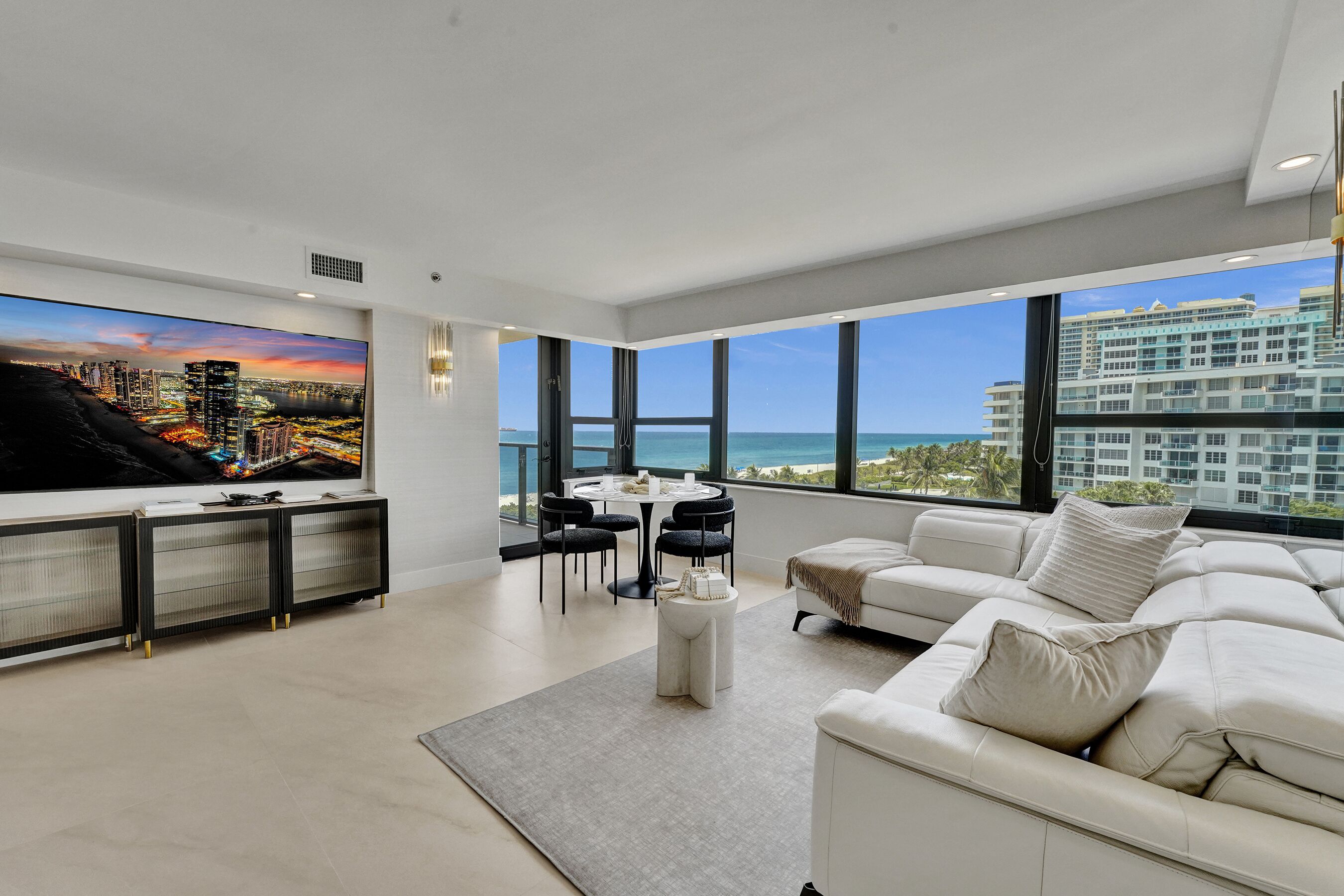 A modern living room with a large TV, a white sofa, a dining table with chairs, and a view of the ocean and buildings.