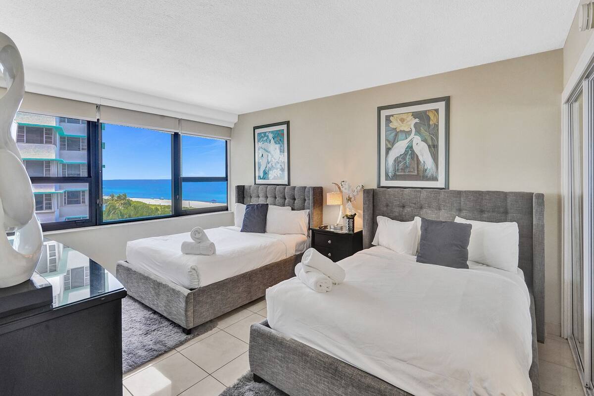 Quarto de hotel com duas camas, uma mesinha de cabeceira e vista para o oceano.